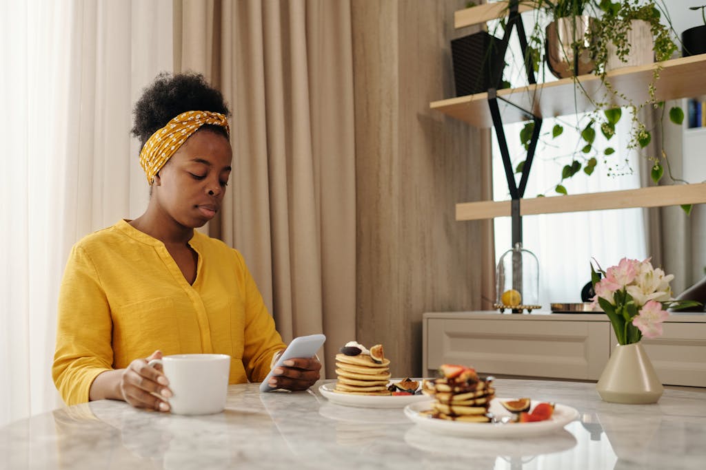 African American woman using smartphone while having pancakes and coffee at a cozy breakfast indoors.