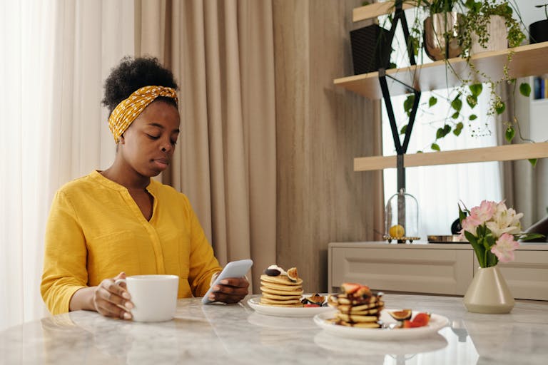African American woman using smartphone while having pancakes and coffee at a cozy breakfast indoors.